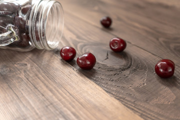 Red juicy cherries scattered from out the glass jar on a brown wooden table from the upper-left corner. Close-up