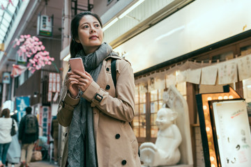 Happy asian woman holding smart phone in front of restaurant. elegant young girl traveler searching direction on online map in cellphone. asian lady visiting local market in shinsekai osaka japan