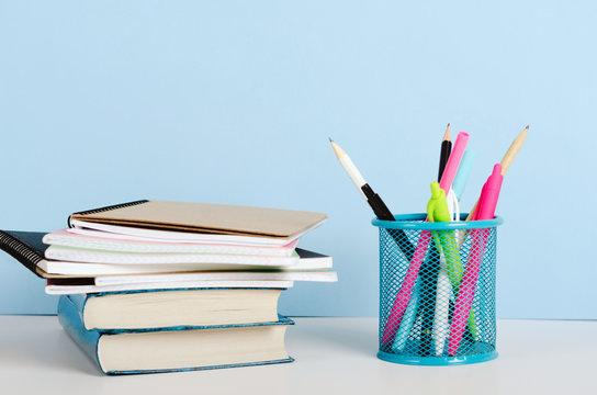 Multi-colored Pencils, Pens In A Stand And Books With Notebooks On A White Table On A Blue Background, Office Workplace. Copy Space.