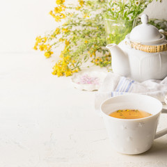 Cup of yellow herbal tea on white table background with teapot and fresh healing herbs and flowers. Herbal medicine. Natural dietary supplement