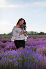 Young, beautiful, elegant woman with long hair and tattoos. Holding and smelling a fragrant bouquet. Girl standing in the field among blooming lavender flowers in the summer in the countryside. 