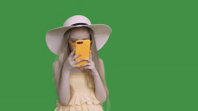 Child girl is browsing he smartphone and surfing ib social media. She is wearing yellow dress, hat and sunglasses. She covers her face with the phone. Keyed green screen alpha channel