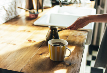 Young woman in grey dress pours natural fresh coffee in a mug in kitchen, morning, hard light