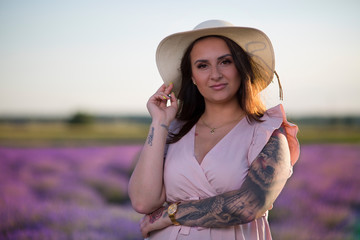 Young, beautiful girl with a hat, long hair and tattoos in a dress. Woman standing in a field of lavender, among blooming, colorful flowers in the countryside in the evening during a summer sunset