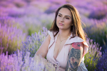 Young, beautiful girl with long hair and tattoos, wearing a pastel dress. Woman sitting in the midst of blooming, colorful flowers in a lavender field in the countryside during a summer sunset.