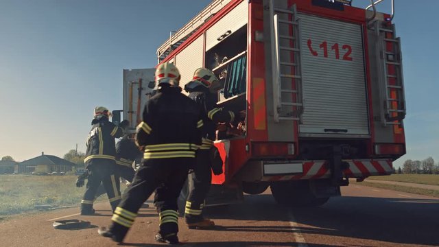 Rescue Team of Firefighters Arrive at the Crash, Catastrophe, Fire Site on their Fire Engine. Firemen Grab their Equipment, Prepare Fire Hoses and Gear from Fire Truck, Rush to Help Injured People.