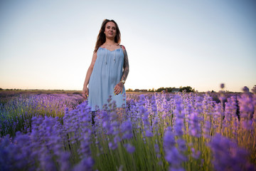 Young, beautiful, pretty girl with tattoos in a dress. Woman standing among blooming colorful flowers in a lavender field during a warm summer sunset.