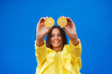 Young Caucasian brunette in yellow blouse holding slices of lemon in front of blue background. Selective focus on lemon.