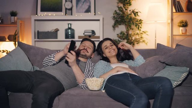 Happy young people man and woman are watching TV laughing and eating popcorn lying on couch at home enjoying leisure time and entertainment in apartment.