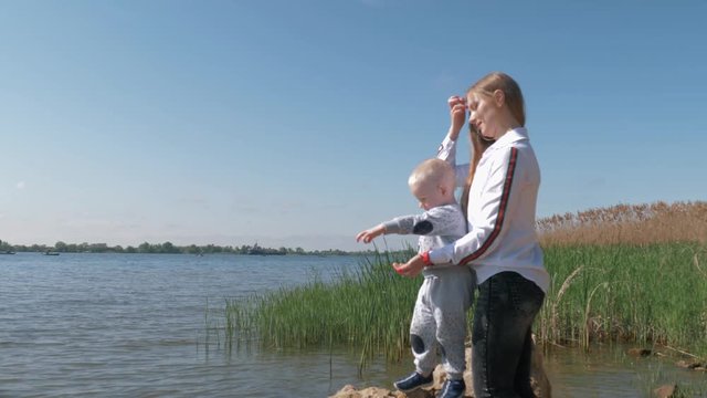 family outdoor recreation, happy woman with baby boy throw food into water for fish in lake during holidays on nature