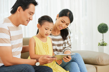 Cute little girl sitting on sofa and watching something on digital tablet together with her parents in the living room at home