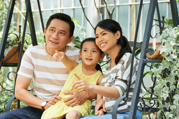 Asian little girl sitting between her mom and dad and pointing at something they sitting on swing together outdoors