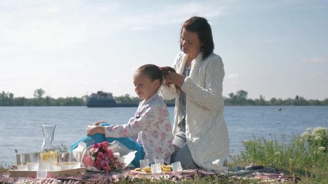 maternal care, happy mom does daughter braid during rest on family picnic on meadow near loch