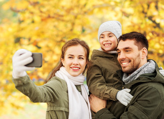 Fototapeta premium family, leisure and people concept - happy mother, father and little daughter taking selfie by smartphone over autumn park background