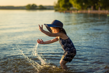 little girl in a hat by the river in summer splashes water