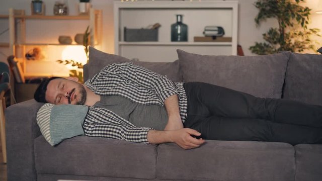 Portrait of person handsome guy napping on sofa at home at night with remote control in hand, TV clicker is falling on floor. People and lifestyle concept.