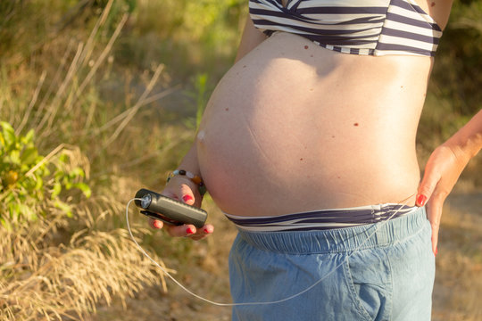 A Diabetic Pregnant Woman With An Insulin Pump In A Forest. She's Wearing A Blue Skirt And A Striped Bikini Top.