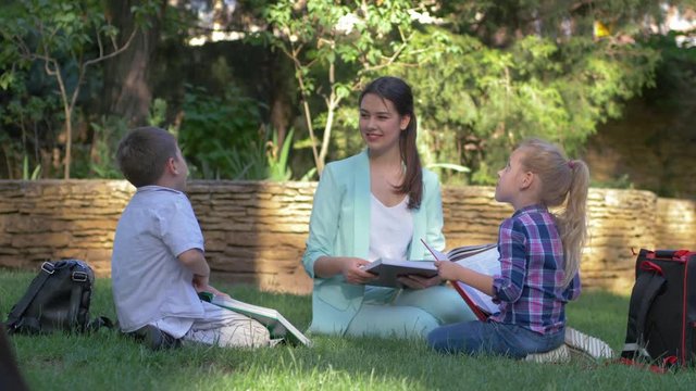 elementary school, happy schoolkids boy and girl with tutor woman read classbook and chat during training on grass outdoors