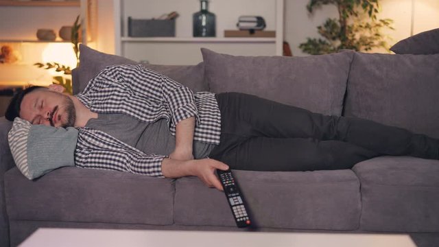 Portrait of tired person bearded man sleeping on couch alone holding TV remote control, clicker is falling on floor. People, lifestyle and casual life concept.