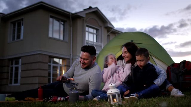 Full shot of happy Caucasian family enjoying picnic in evening in back yard of their large house, father roasting sausage on gas cooker, while loving mother is hugging and laughing with children