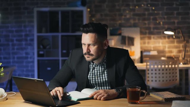 Bearded guy is using laptop at night in office and making notes in notebook sitting at desk working late. Workaholics, workplace and business concept.
