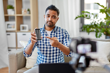 blogging, videoblog and people concept - sad disappointed indian male blogger with camera recording video review of smartphone at home