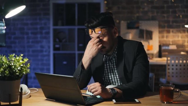 Portrait of tired office worker handsome guy in glasses using laptop yawning working in office late at night. Workaholics, overwork and business concept.