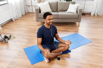 fitness, meditation and healthy lifestyle concept - indian man in earphones listening to music on smartphone and meditating in lotus pose at home