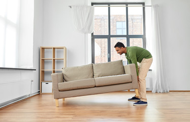 accommodation, furniture and interior concept - happy indian man moving sofa at home