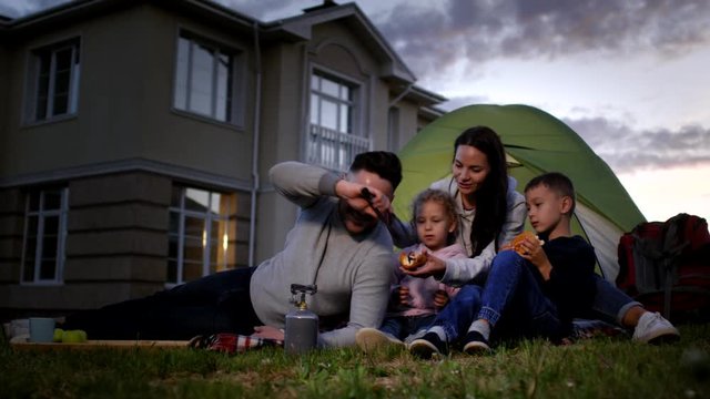 Full shot of smiling middle-aged Caucasian man making hotdogs for his family on lawn in front of suburban home, putting roasted sausage in bread roll, then wife adding ketchup while kids watch