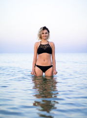 Obraz premium female with curly hairs in black swimsuit is standing in the sea, sunset time