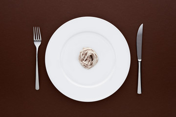 Small portion of tagliatelle pasta on round white plate with fork and knife on dark brown tablecloth. Concept of dieting and healthy eating.