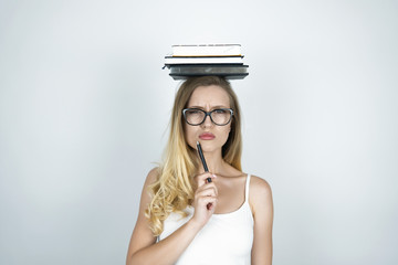 serious young woman in glasses holding pen in her hand and books on her head isolated white...