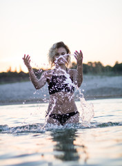 curly hair girl in black swimsuit standing in sea water and sprying water, sunset on the background
