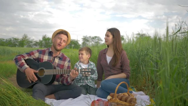 fun family vacation, merry male plays musical instrument while woman with child sing and clap while relaxing on picnic outdoors in green field close-up