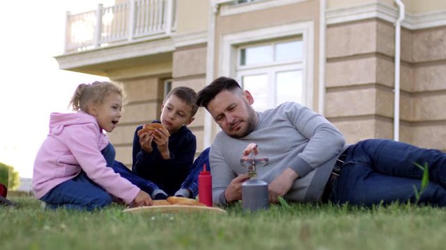 Medium shot of 40-something Caucasian father and two children lying on grass in back yard of suburban apartment complex, eating hotdogs, roasting sausages on portable gas cooker and chatting