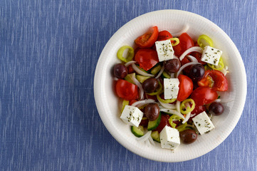 Traditional greek salad in white plate on blue matting with empty space.
