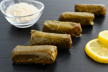 Delicious stuffed grape leaves (the traditional dolma of the mediterranean cuisine) on black dish with lemon slices and small glass bowl of raw rice
