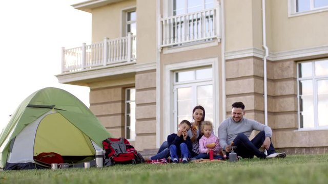Full shot of happy Caucasian mom and dad enjoying picnic with kids on green lawn in back yard, with camping tent and backpacks nearby, roasting sausages on portable gas cooker and eating hotdogs