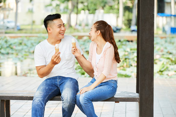 Asian young couple in jeans sitting on bench eating ice cream and laughing while resting in the park