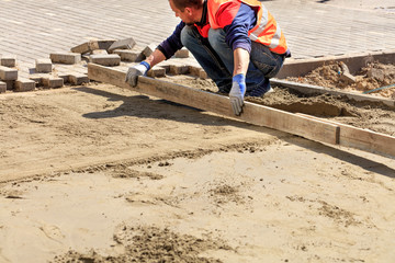 Worker aligns the sand base with a wooden board for laying paving slabs on the sidewalk.