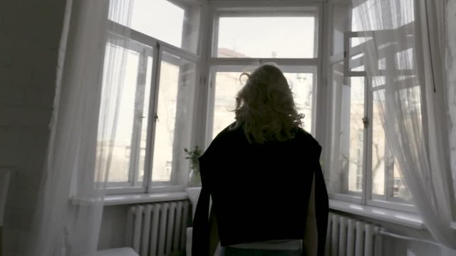 Rear view of a beautiful slim blonde at home by the window. Action. Back view of a young girl with black cardigan on her shoulders walking towards the window.