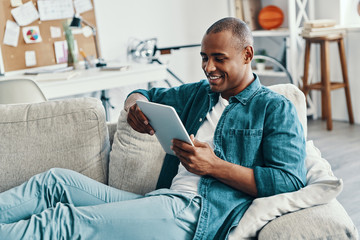 Totally carefree. Handsome young African man using digital tablet and smiling while sitting indoors