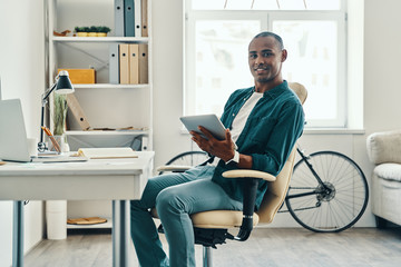 Putting ideas into something real. Handsome young African man in shirt working using digital tablet while sitting in the office