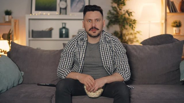 Shocked guy is watching TV with attention and eating popcorn looking at screen sitting on couch in apartment alone. Modern lifestyle and people concept.