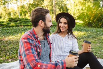 Image of pleased couple man and womandrinking takeaway coffee while resting in green park