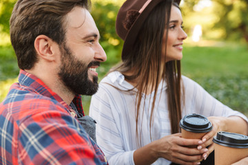 Image of attractive couple man and woman drinking takeaway coffee while resting in green park