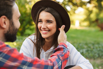 Image of caucasian couple man and woman smiling and looking at each other while resting in green park