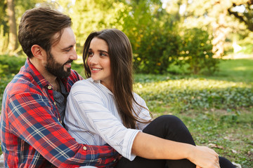 Fototapeta premium Image of optimistic couple man and woman hugging and looking at each other while resting in green park