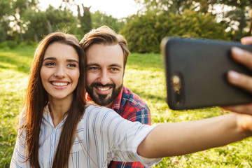 Image of happy couple man and woman taking selfie photo on cellphone while resting in green park